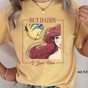 May include: Mustard-colored t-shirt featuring a graphic of Ariel and Flounder from The Little Mermaid, with the text "BUT DADDY I Love Him." The design is framed in a brown rectangle. The shirt is a comfortable, casual style.