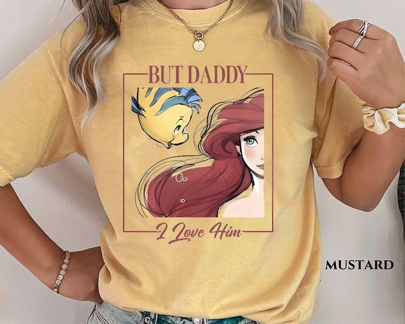 May include: Mustard-colored t-shirt featuring a graphic of Ariel and Flounder from The Little Mermaid, with the text "BUT DADDY I Love Him." The design is framed in a brown rectangle. The shirt is a comfortable, casual style.
