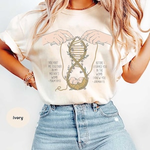 Puede incluir: Camiseta de color marfil con un dibujo lineal de manos tejiendo una estructura similar al ADN con un bebé en una cuna. La camiseta incluye el texto "YOU KNIT ME TOGETHER IN MY MOTHER'S WOMB - PSALM 139:13" y "BEFORE I FORMED YOU IN THE WOMB I KNEW YOU - JEREMIAH 1:5".