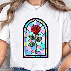 May include: White t-shirt with a stained glass rose design. The graphic shows a red rose with green leaves, within a colourful, arched window frame. The design is centered on the shirt.