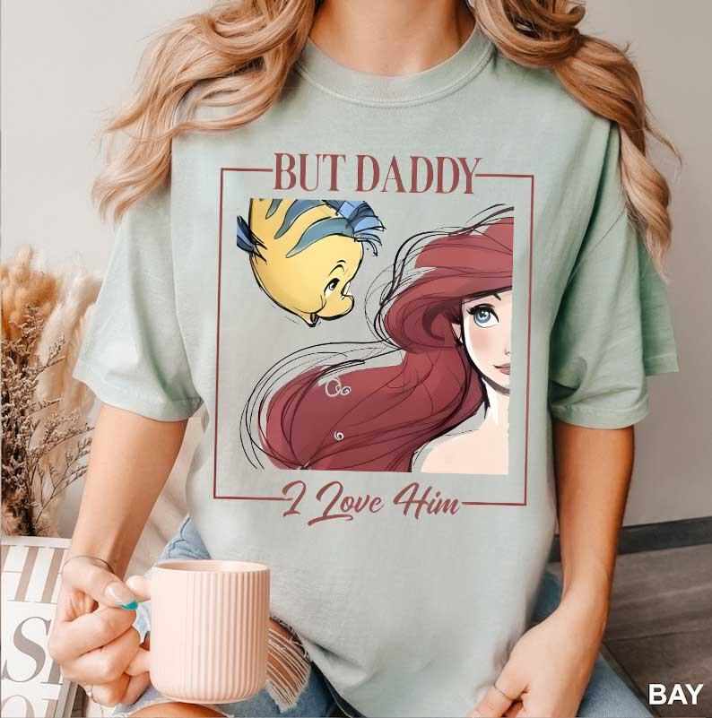 May include: Sage green t-shirt featuring a graphic of Ariel and Flounder from The Little Mermaid, with the text "BUT DADDY - I Love Him" in a rectangular frame. The design includes red, yellow, and blue colors.