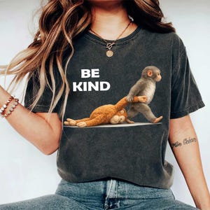 Pode incluir: T-shirt cinza escuro com as palavras "BE KIND" em branco. A camisa apresenta um gráfico de dois macacos, um puxando o outro. A camisa é feita de um material macio.
