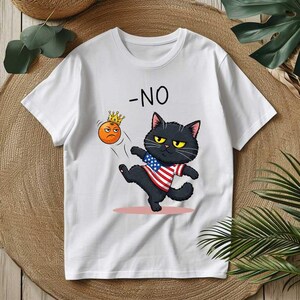 May include: White t-shirt featuring a cartoon black cat kicking an orange emoji wearing a crown. The cat is wearing a shirt with an American flag design. The word "-NO" is printed above the cat.
