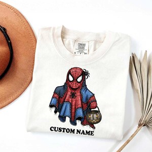以下が含まれることがあります： ハロウィーンをテーマにしたスパイダーマンのデザインが施された、クリーム色のTシャツ。グラフィックは、カボチャのバケツを持っている幽霊に扮したスパイダーマンを示しています。「CUSTOM NAME」の文字が下に印刷されています。シャツにはComfort Colorsのタグが付いています。