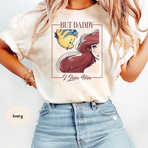 May include: Ivory-colored t-shirt featuring a graphic of Ariel and Flounder from The Little Mermaid, with the text "BUT DADDY - I Love Him" in a rectangular frame. The shirt is a light color.