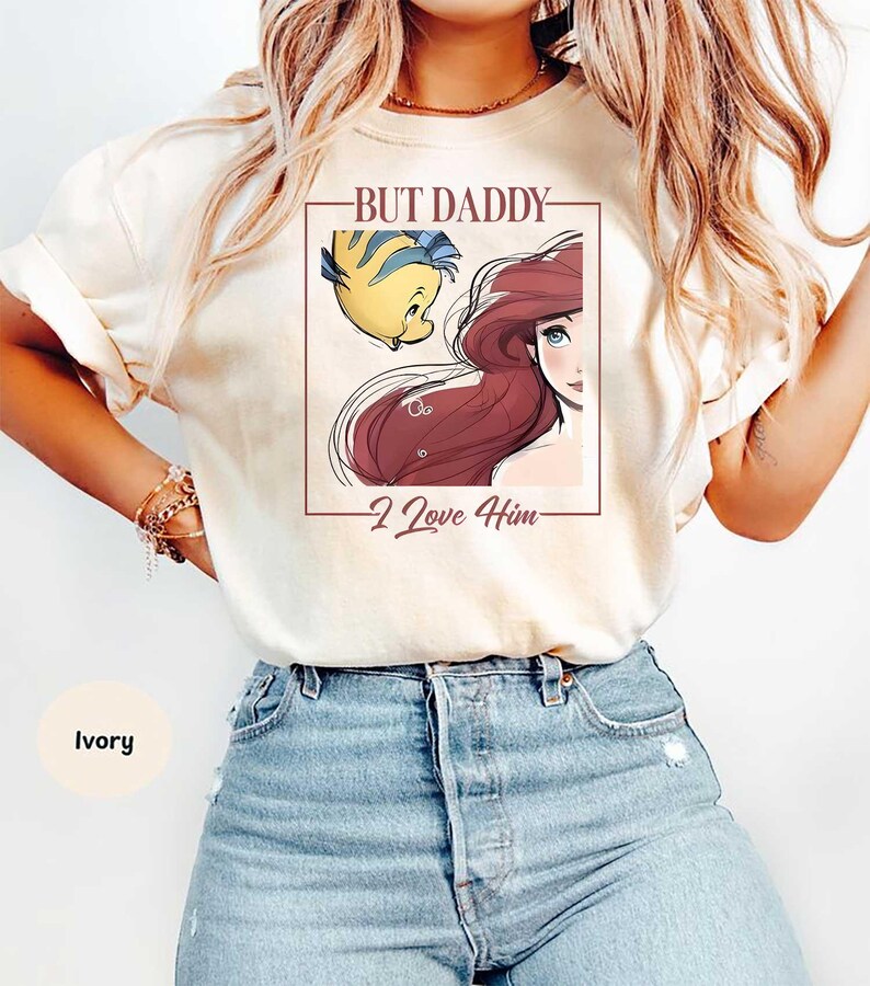 May include: Ivory-colored t-shirt featuring a graphic of Ariel and Flounder from The Little Mermaid, with the text "BUT DADDY - I Love Him" in a rectangular frame. The shirt is a light color.