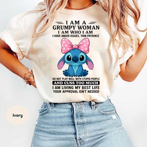 Könnte beinhalten: Elfenbeinfarbenes T-Shirt mit einer Cartoonfigur und einer rosa Schleife. Das Shirt trägt den Text "I AM A GRUMPY WOMAN I AM WHO I AM I HAVE ANGER ISSUES, THIN PATIENCE. DO NOT PLAY WELL WITH STUPID PEOPLE AND CUSS TOO MUCH I AM LIVING MY BEST LIFE YOUR APPROVAL ISN'T NEEDED."