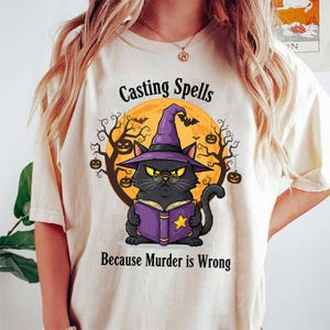 May include: Cream-colored t-shirt featuring a black cat wearing a witch hat and glasses, holding a purple book. The graphic includes the text "Casting Spells Because Murder is Wrong" with a full moon and Halloween-themed elements.