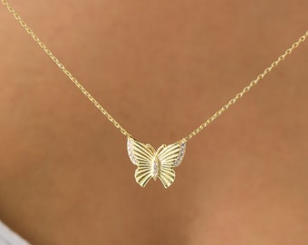 May include: A gold-colored butterfly pendant necklace. The butterfly has detailed wing patterns and a small gem in the center. The necklace chain is delicate and hangs on a person.