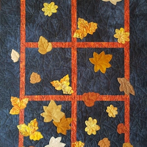 Peut inclure: Une courtepointe avec un fond bleu foncé et un cadre rouge. La courtepointe présente un motif de feuilles jaunes, orange et brunes éparpillées sur la surface.