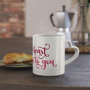 Peut inclure: Tasse en céramique blanche avec une anse en forme de cœur. La tasse porte l'inscription "my heart is to you" en script rouge. Un croissant est posé sur une assiette verte à gauche de la tasse.