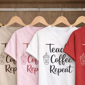Könnte beinhalten: Eine Reihe von T-Shirts in Schwarz, Beige, Rosa, Weiß und Rot, die an Holzkleiderbügeln hängen. Die Shirts zeigen den Text "Teach Coffee Repeat" mit einer Kaffeetassen-Grafik. Die Shirts befinden sich in einem Holzschrank.