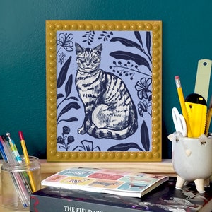 A framed illustration of a cat sitting in a blue floral pattern. The frame is yellow with a beaded border. Surrounded by stationary, in front of dark green wall.