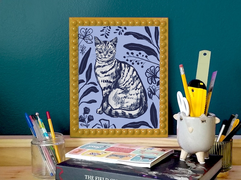 A framed illustration of a cat sitting in a blue floral pattern. The frame is yellow with a beaded border. Surrounded by stationary, in front of dark green wall.