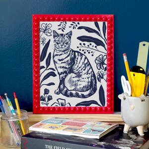 A dark blue illustration of a cat  with flowers and leaves surrounding it. Background of the illustration is white. The portrait is in a red beaded frame, in front of dark blue wall and standing on wooden surface, surrounded by stationery.