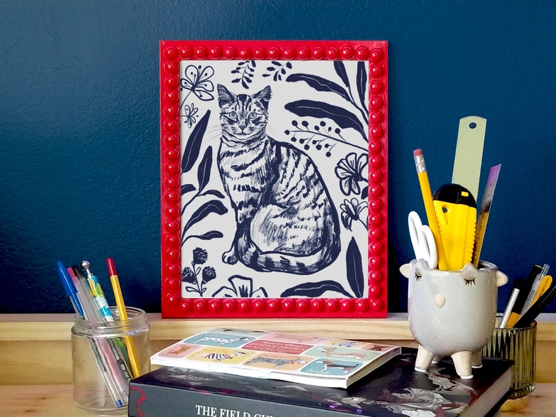 A dark blue illustration of a cat  with flowers and leaves surrounding it. Background of the illustration is white. The portrait is in a red beaded frame, in front of dark blue wall and standing on wooden surface, surrounded by stationery.