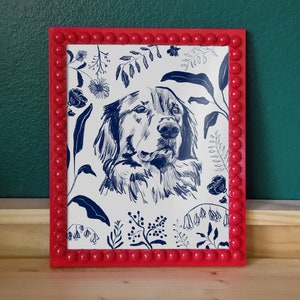 A dark blue illustration of a dog's head with flowers and leaves surrounding it. Background of the illustration is white. The portrait is in a red beaded frame, in front of dark green wall and standing on wooden surface.
