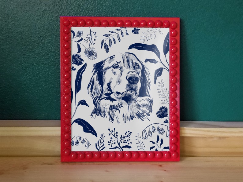 A dark blue illustration of a dog's head with flowers and leaves surrounding it. Background of the illustration is white. The portrait is in a red beaded frame, in front of dark green wall and standing on wooden surface.