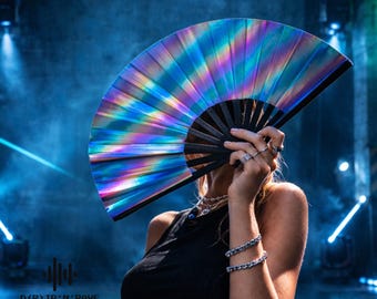 May include: A handheld folding fan featuring a reflective, iridescent surface that displays a spectrum of rainbow colors. The fan is constructed with black ribs and a black frame. The text D(R)IP'N'RAVE is printed in the lower left corner.