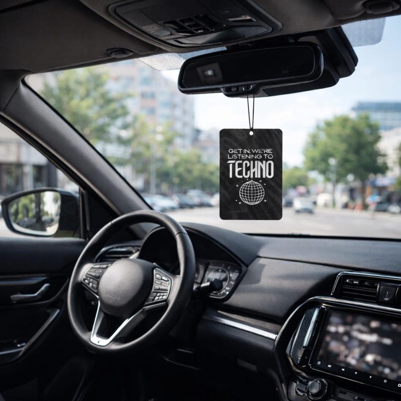 May include: Black rectangular car air freshener with the text "GET IN WE'RE LISTENING TO TECHNO" and a globe design. The air freshener hangs from the rearview mirror inside a car, with the steering wheel and dashboard visible.