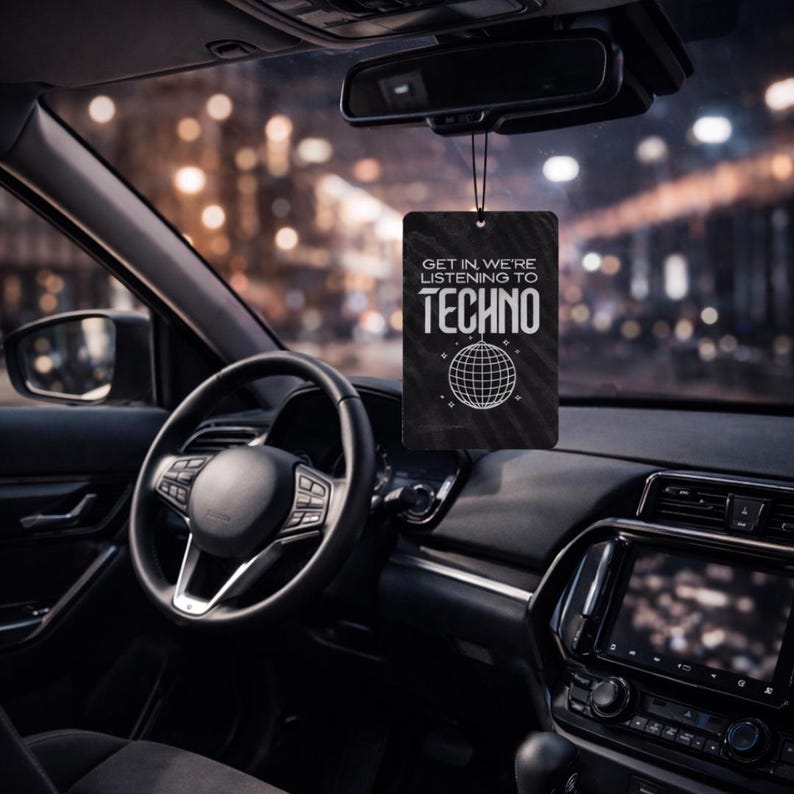 May include: Black car air freshener with the text "GET IN WE'RE LISTENING TO TECHNO" and a disco ball graphic. The air freshener hangs from the rearview mirror in a car interior with a blurred city background.