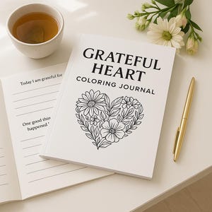 May include: A white "Grateful Heart Coloring Journal" with a floral heart design, a gold pen, and a cup of tea. The journal is open to a page with prompts. White flowers are in the background.