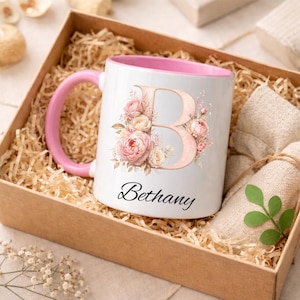 May include: White ceramic mug with a pink interior and handle, featuring a floral "B" design and the name "Bethany" in script. The mug is presented in a gift box with decorative paper and a wrapped cloth.