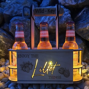 May include: Wooden six-pack bottle carrier with the text "Enjoy the Lillet Things" and a graphic of raspberries. The carrier is holding three bottles of Lillet.