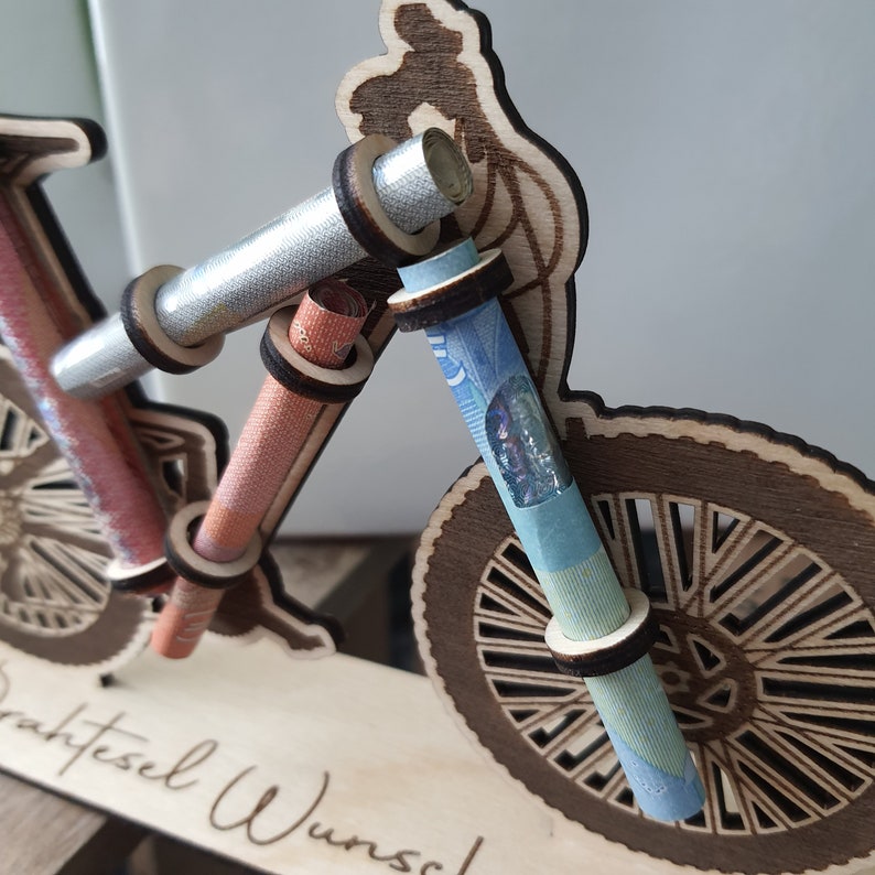 May include: Wooden bicycle money holder with rolled up bills inserted into the handlebars. The base of the holder has the text "Gutesel Wunsch" engraved on it.