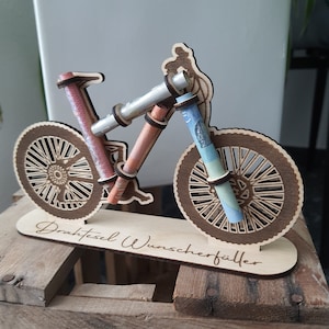 May include: Wooden bicycle money holder with a person riding on it. The bicycle has a brown base and the words "Drahtesel Wunschersch&ouml;pfer" are written on it.