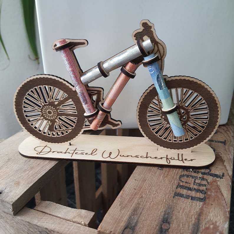 May include: A wooden bicycle money holder with the text "Drahtesel Wunscherschfiller" on the base. The bicycle has a silver, pink, and blue rolled up bills in the handlebars and wheels.