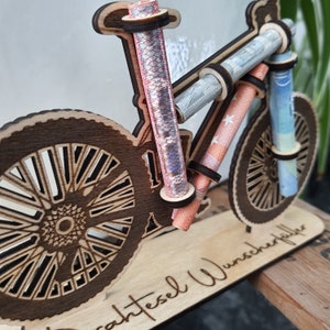 May include: A wooden bicycle money holder with a brown finish. The bicycle has a brown base with the text "Sachtesel Wunschkoffer" engraved on it. The bicycle has two wheels and a handlebar with a place to hold money.