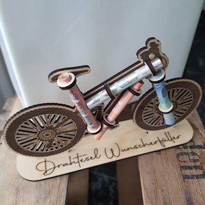 May include: A wooden bicycle money holder with the text "Drahtesel Wunscherschfiller" on the base. The bicycle is decorated with rolled up bills of money.