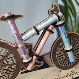 May include: A wooden bicycle model with rolled up bills of money inserted into the handlebars and wheels. The bicycle is on a wooden base with the word "cherfille" written on it.