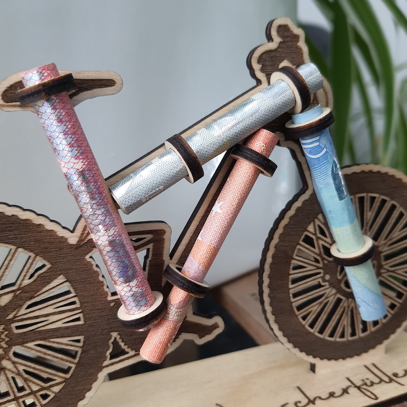 May include: A wooden bicycle model with rolled up bills of money inserted into the handlebars and wheels. The bicycle is on a wooden base with the word "cherfille" written on it.