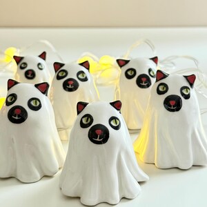 Könnte beinhalten: Eine Sammlung weißer Keramik-Katzenfiguren, die als Geister verkleidet sind. Jede Figur hat eine schwarze Nase, grüne Augen und rot gefärbte Ohren. Eine Lichterkette mit gelben Lichtern im Hintergrund erzeugt ein Halloween-Thema.