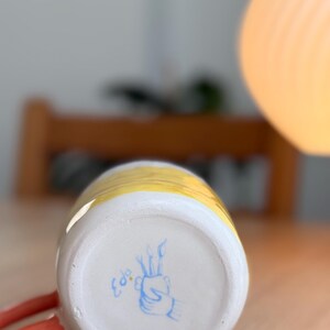 K&ouml;nnte beinhalten: Eine gelbe und wei&szlig;e Keramiktasse mit einem roten Henkel. Der Boden der Tasse hat ein handgemaltes Design in Blau mit dem Wort "Colo" in Blau.