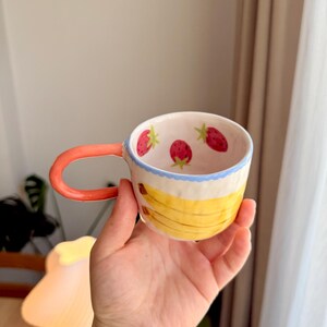 K&ouml;nnte beinhalten: Eine Keramiktasse mit rotem Henkel und wei&szlig;em Hintergrund. Die Tasse ist mit gelben Bananen und roten Erdbeeren verziert.