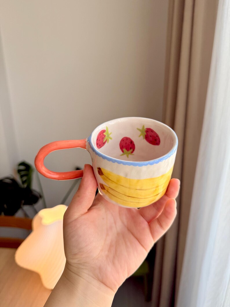 K&ouml;nnte beinhalten: Eine Keramiktasse mit rotem Henkel und wei&szlig;em Hintergrund. Die Tasse ist mit gelben Bananen und roten Erdbeeren verziert.