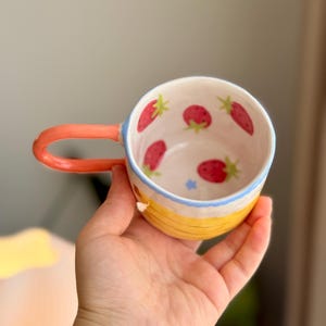 K&ouml;nnte beinhalten: Eine gelbe Keramiktasse mit Erdbeer-Design. Die Tasse hat einen hellblauen Rand und einen hellorangenen Henkel.