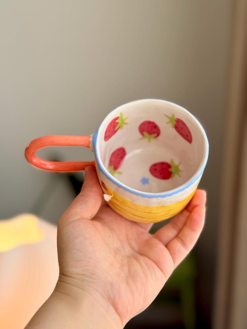 K&ouml;nnte beinhalten: Eine gelbe Keramiktasse mit Erdbeer-Design. Die Tasse hat einen hellblauen Rand und einen hellorangenen Henkel.