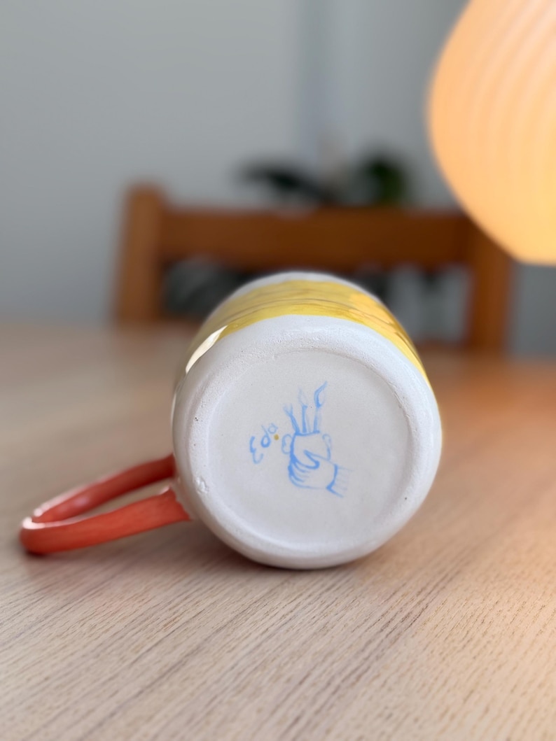 K&ouml;nnte beinhalten: Eine gelbe und wei&szlig;e Keramiktasse mit einem roten Henkel. Der Boden der Tasse hat ein handgemaltes Design in Blau mit dem Wort "Colo" in Blau.