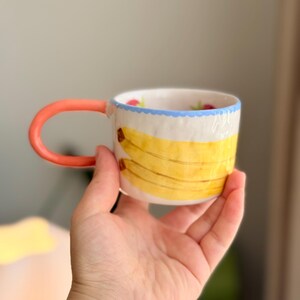 K&ouml;nnte beinhalten: Eine wei&szlig;e Keramiktasse mit einem roten Henkel und einem blauen und wei&szlig;en Rand. Die Tasse hat ein gelbes Bananenmuster mit gr&uuml;nen Bl&auml;ttern und roten Beeren.