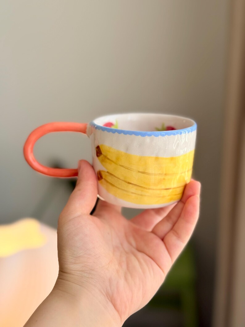 K&ouml;nnte beinhalten: Eine wei&szlig;e Keramiktasse mit einem roten Henkel und einem blauen und wei&szlig;en Rand. Die Tasse hat ein gelbes Bananenmuster mit gr&uuml;nen Bl&auml;ttern und roten Beeren.