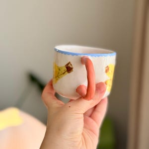 K&ouml;nnte beinhalten: Eine wei&szlig;e Keramiktasse mit einem hellblauen Rand und einem rosafarbenen Henkel. Die Tasse hat ein gelbes und braunes Design auf der Vorderseite.