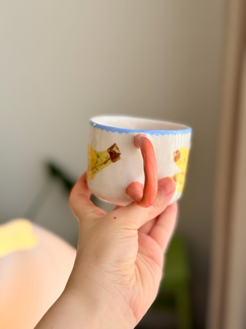 K&ouml;nnte beinhalten: Eine wei&szlig;e Keramiktasse mit einem hellblauen Rand und einem rosafarbenen Henkel. Die Tasse hat ein gelbes und braunes Design auf der Vorderseite.