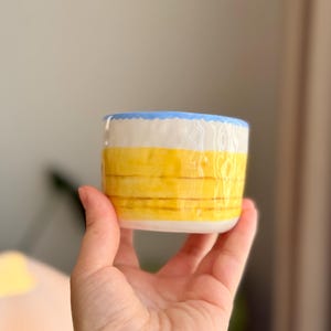 K&ouml;nnte beinhalten: Eine kleine, gelbe Keramiktasse mit blauem Rand. Die Tasse hat ein gestreiftes Muster und wird in einer Hand gehalten.