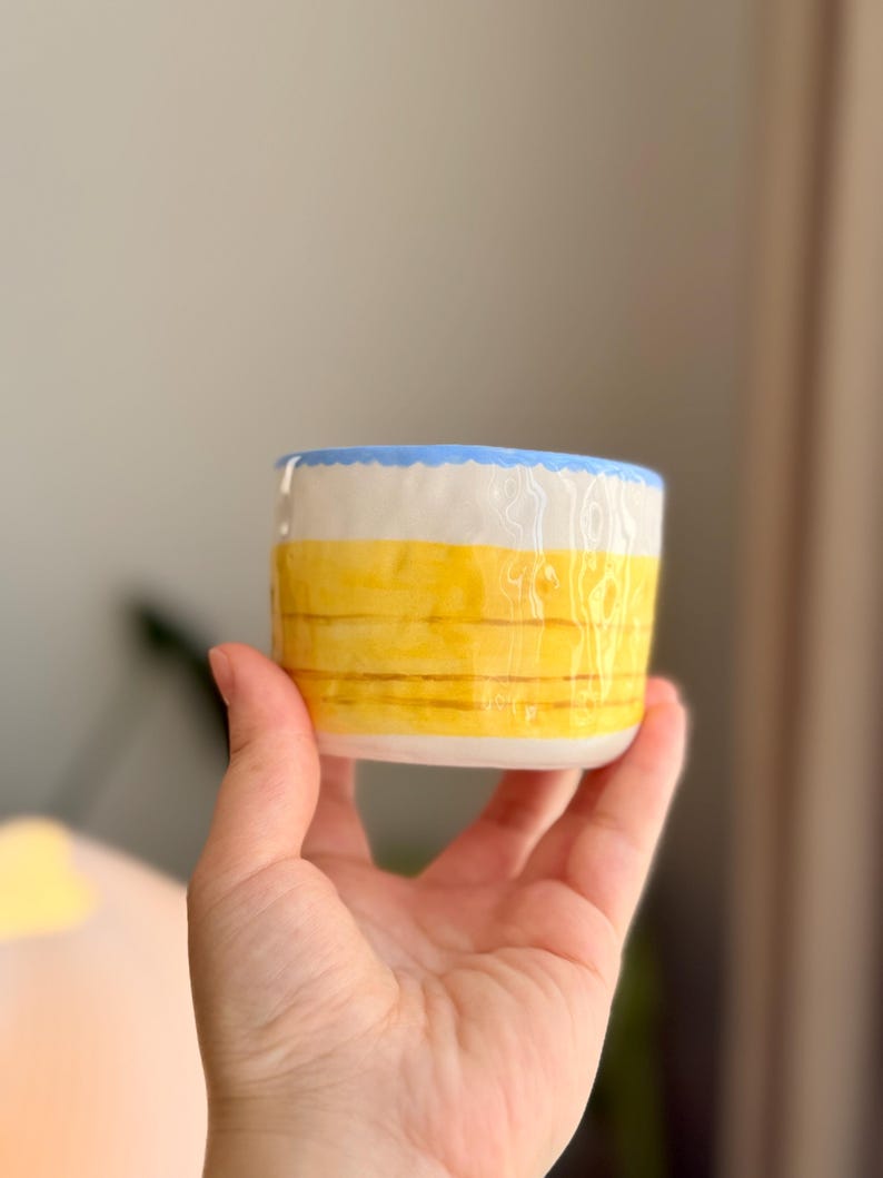K&ouml;nnte beinhalten: Eine kleine, gelbe Keramiktasse mit blauem Rand. Die Tasse hat ein gestreiftes Muster und wird in einer Hand gehalten.