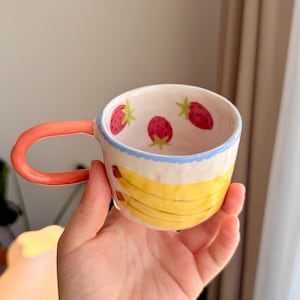 K&ouml;nnte beinhalten: Eine Keramiktasse mit einem hellblauen Rand und einem rosafarbenen Henkel. Die Tasse ist mit handbemalten gelben Bananen und roten Erdbeeren verziert.
