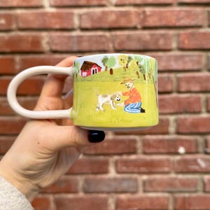 May include: A white ceramic mug with a handle features a hand-painted illustration of a person and a dog in a green field. The scene includes a small red house and trees.